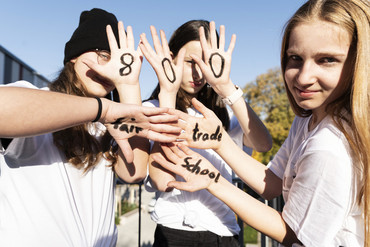 3 Schülerinnen halten ihre Handflächen in die Kamera, auf denen die Zahlen 8, 0 und 0 sowie die Worte "fair", "trade" und "school" stehen 